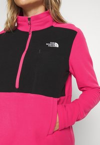 Fleecejacka med halvdragkedja, svart överdel och rosa underdel samt en bröstficka. Logotypen visas på vänster bröst.