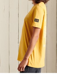 Camiseta de algodón amarilla con mangas cortas, presenta un diseño gráfico en la parte delantera y una etiqueta de tela en la manga. Corte recto.