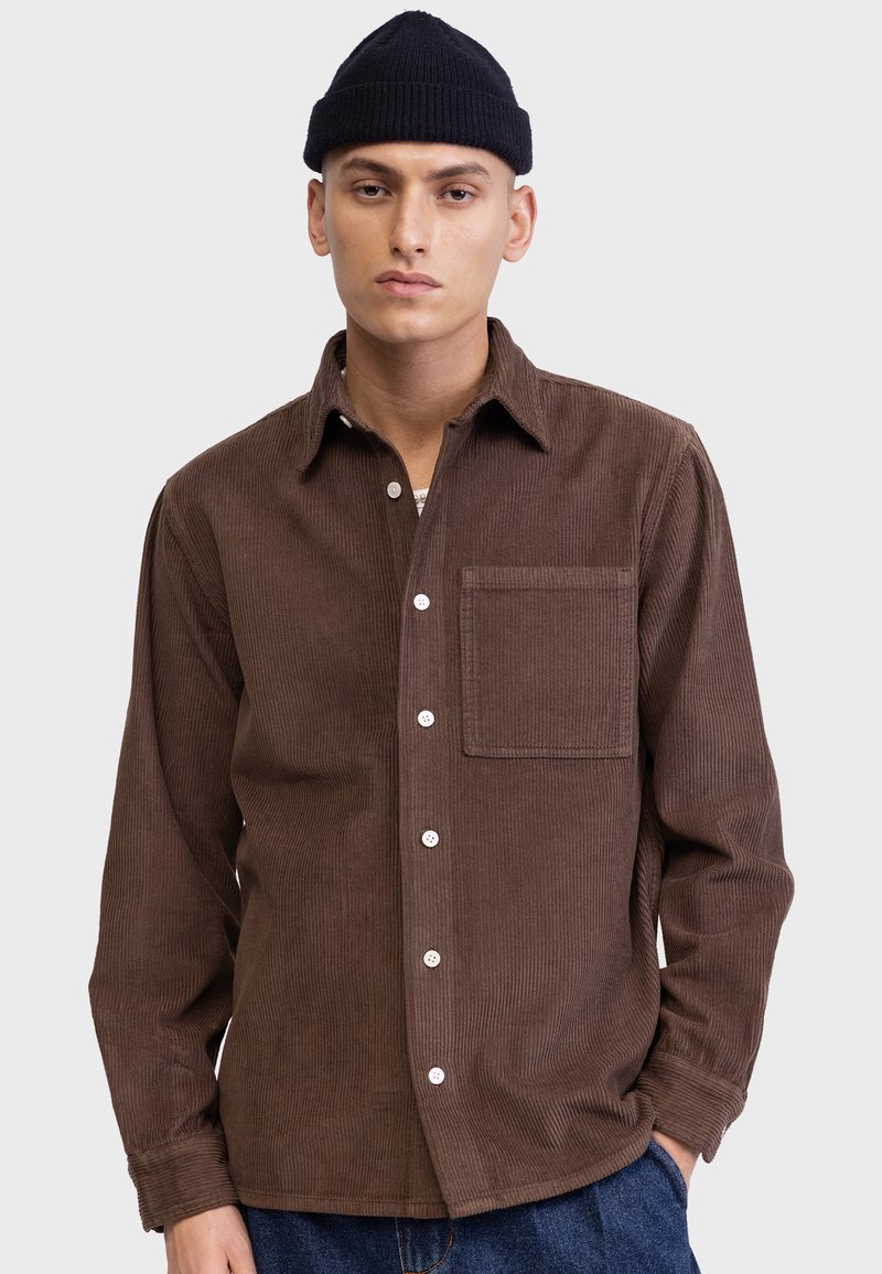 Camisa de pana marrón con frente abotonado, un bolsillo en el pecho y botones blancos. Manga larga, corte relajado, cuello estructurado.