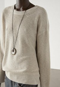 Pull en tricot beige à la coupe ample et au décolleté large. Manches longues, tissu texturé et ourlet côtelé. Assorti à un collier avec un grand pendentif circulaire.
