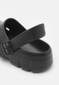 Sandal in gomma nera con una larga cinghia e fibbia regolabile. La suola è spessa, con una finitura testurizzata e una piattaforma rialzata.