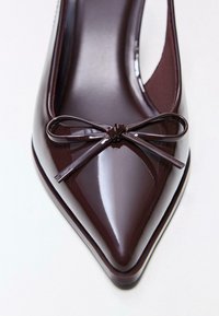 Escarpin en cuir verni bordeaux foncé avec un bout pointu, ornée d'un petit nœud sur le dessus et une finition brillante.