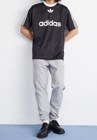 Černé tričko Adidas s bílým logem a pruhy, spárované se světle šedými kalhotami se zipy a elastickými manžetami, nošeno s černými botami.