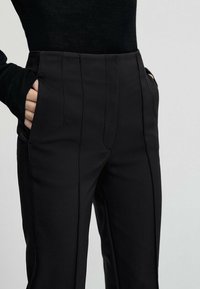 Black tailored trousers with a smooth texture, featuring side pockets and subtle pleats at the front for a refined look.