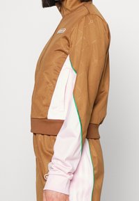 Brown athletic jacket with a high collar, pink sleeves, green piping, and all-over geometric pattern. Features a front zipper and logo patch.