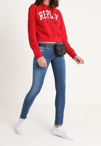 Sudadera recortada roja con letras "REPLAY", vaqueros ajustados azules, zapatillas blancas y un pequeño bolso negro de cinturón. Tejido suave, diseño casual.