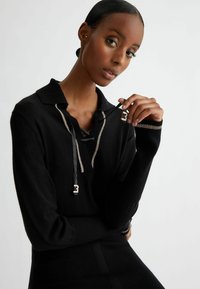 Zwarte pullover trui met een kraag, zilveren accenten en zwarte linten. Heeft een opvallende logo-detail nabij de hals.