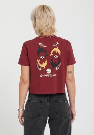 Personne aux cheveux blonds courts portant un haut court bordeaux avec des dauphins, des yeux, des étoiles et le texte "Cosmic Rider" dans le dos.