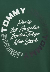 Πράσινο ύφασμα με κυκλικό κείμενο "Tommy Jeans" που περιβάλλει τα ονόματα των πόλεων Παρίσι, Λος Άντζελες, Λονδίνο, Τόκιο και Νέα Υόρκη σε λευκή γραμματοσειρά καλλιγραφίας.