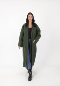 Lange, oversized groene jas van dikke stof, met brede mouwen en voorzakken, gecombineerd met blauwe jeans en zwarte enkellaarsjes.