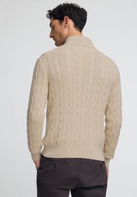 Beige kabelstickad tröja med ribbad krage och manschetter. Har texturerade mönster och en figurnära silhuett, bärs tillsammans med mörka byxor.