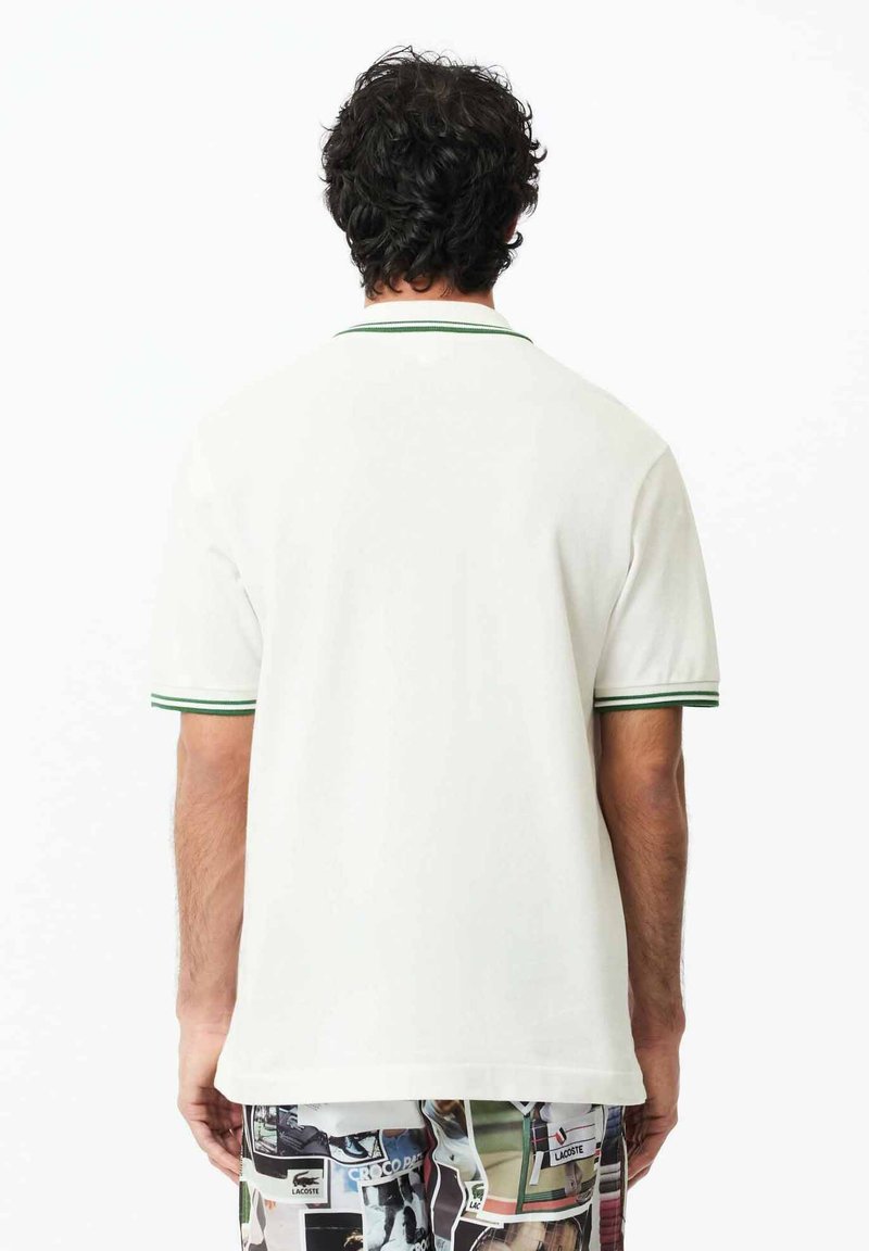 Polo blanc avec un col et des manches à rayures vertes. Tissu lisse, coupe classique. La vue arrière montre un design simple et épuré.