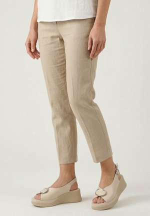 Frau trägt beige kurze Hosen und beige Keilsandalen mit silberner Schnalle, steht vor hellem Hintergrund.