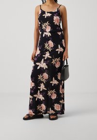 Robe maxi noire à fleurs avec fines bretelles, ornée de fleurs rose clair et violettes, tissu fluide, associée à des sandales plates noires et un petit sac.