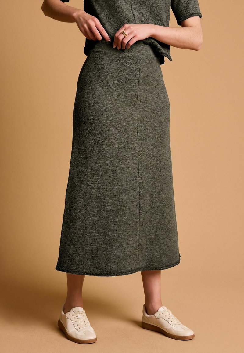 Jupe olive verte mi-longue en tissu texturé, avec une taille cintrée et un détail subtil à l'ourlet, associée à des baskets beiges.