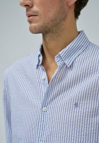 Camisa de botón a rayas azul y blanca, de tela de algodón, con un cuello clásico y un pequeño emblema bordado en el pecho.