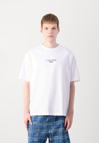 Camiseta blanca de algodón con cuello redondo, mangas cortas y el logo negro "Calvin Klein JEANS" en el pecho. El modelo lleva pantalones azules con estampado.