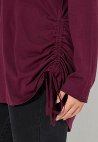 Bordeaux farbenes Langarmoberteil mit Raffung an der Seite und Bindebändern am Saum. Weicher Stoff mit glatter Textur. Getragen über dunklen Jeans.