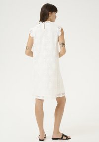 Rochie albă din dantelă cu un model floral, mâneci fluture, detaliu cu deschidere la spate, de lungime până la genunchi, asortată cu sandale negre cu talpă joasă.