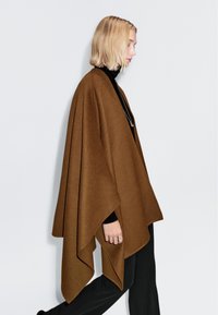 Brun poncho gjort av mjuk tyg, med en lös, hängande design, breda ärmar och en slät textur, bärs över en svart polotröja.