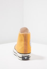Žluté plátěné vysoké tenisky s texturovaným povrchem, bílou gumovou podrážkou, černými akcenty a nášivkou loga Converse na vnější části paty.