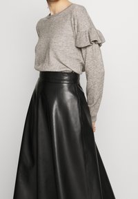 Femme portant un pull gris à manches volantées associé à une jupe en cuir noire taille haute en forme A, sur fond uni.