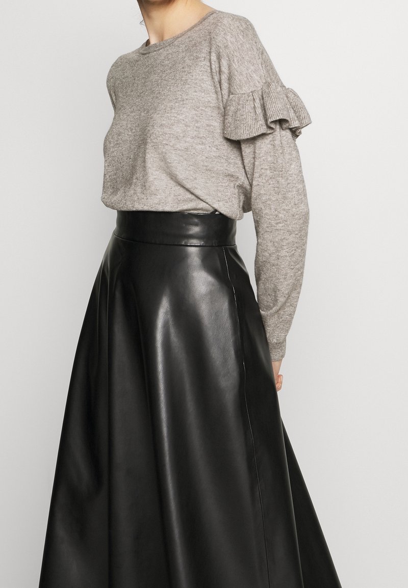 Femme portant un pull gris à manches volantées associé à une jupe en cuir noire taille haute en forme A, sur fond uni.