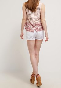 Vaaleanpunainen hihaton paita, jossa on pilkkuja ja kuvioitu helma, yhdistettynä valkoisiin shortseihin ja ruskeisiin korkokenkiin.