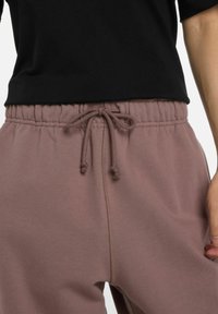 Close-up van een persoon in mauve joggingsbroek met een gestrikt koord en een zwart shirt, hand rustend naast hun lichaam.