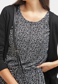 Schwarze Strickjacke über einem gemusterten schwarz-weißen Kleid. Das Kleid hat einen strukturierten, abstrakten Druck und einen Rundhalsausschnitt.