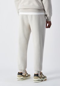 Pantalons de survêtement gris clair en tissu doux, jambe fuselée, taille élastique. Portés avec des baskets beige et noires avec des accents oranges.