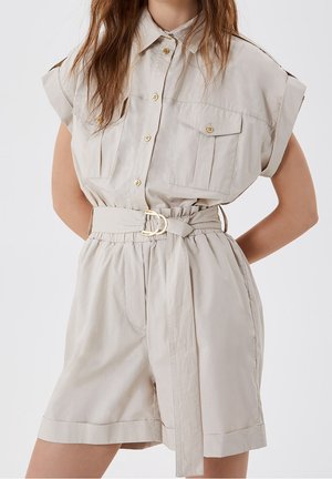 Femme portant une combinaison beige à manches courtes ceinturée, avec boutons sur le devant et poches à rabat sur la poitrine, debout les mains dans le dos.