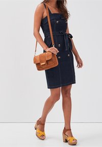 Džínové šaty s upnutým živůtkem, knoflíky a zavazovacím detailem v pase. Kombinované s hnědou semišovou kabelkou přes rameno a žlutými sandály na podpatku.