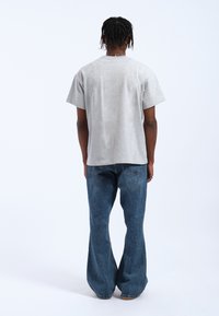 Camiseta gris de manga corta con un ajuste holgado, combinada con unos pantalones vaqueros acampanados azules que presentan una textura desgastada y bolsillos traseros, vistos desde la parte de atrás.