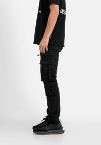 Zwarte cargo broek met meerdere ritszakken, slim fit, gecombineerd met zwarte sneakers. Het T-shirt heeft een contrasterend logo op de borst.