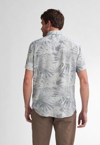 Camicia a maniche corte in tessuto grigio chiaro con un motivo a foglie di palma, con orlo dritto e colletto standard. Indossata con pantaloni beige.