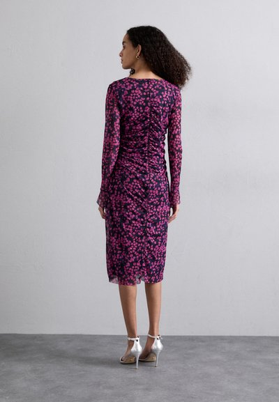 Robe florale violette avec des manches longues, des détails froncés et une longueur jusqu'au genou. Portée avec des sandales à talons hauts argentées. Texture de tissu lisse.