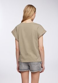 Camicia khaki a maniche corte con risvolti e rifiniture dorate, abbinata a pantaloni corti in denim grigio con orli sfilacciati. Tessuto morbido e vestibilità rilassata.
