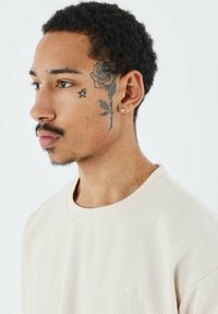 Sudadera de punto de color crema con un patrón texturizado, cuello redondo y pequeño logotipo bordado en el pecho. Vista lateral del modelo con tatuajes.