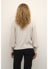 Beige túlméretezett pulóver, struktúrált felülettel, denevérujjakkal és bordás szegéllyel. Fekete nadrággal viselve, hátulról nézve.