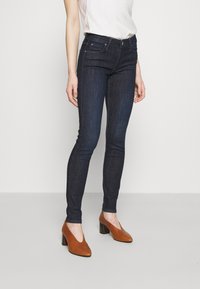 Jeans ajustados de denim en un tono azul oscuro, con cinco bolsillos, costuras sutiles y una cintura de altura media, combinados con zapatos de tacón marrones.