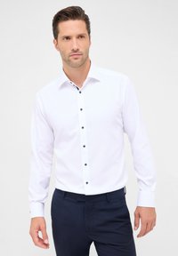 Witte blouse met lange mouwen en een button-down kraag, voorzien van donkerblauwe knopen en accentdetails langs de kraag. Gestyled met marineblauwe broeken.