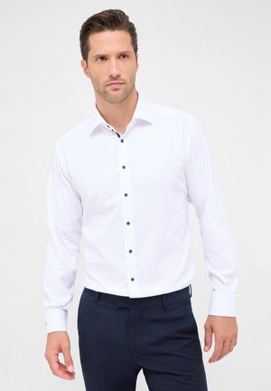 Weißes Langarmhemd mit Button-Down-Kragen, ausgestattet mit dunkelblauen Knöpfen und Akzentdetails entlang des Kragens. Kombiniert mit marineblauen Hosen.