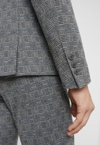 Grijze gestreepte pak met een gestructureerde stof met een ruitpatroon, drieknoopmanchetten detail en bijpassende broek in een close-up weergave.