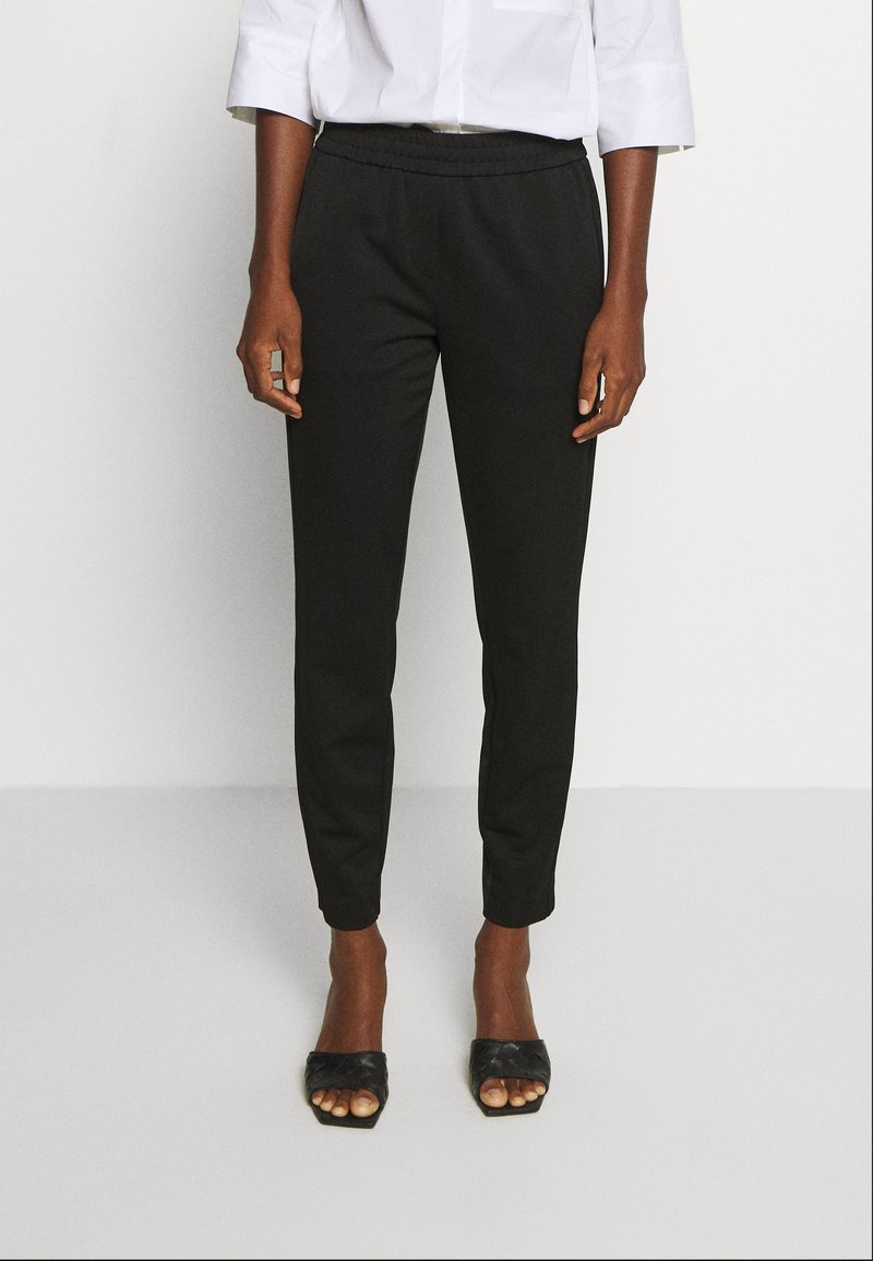 Pantalons noirs ajustés avec une taille élastique, texture lisse, longueur cheville, associés à des sandales ouvertes à bout tissé noires.