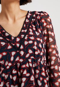 Kukka-kuosiset mekko tummansinisellä taustalla, jossa on V-mallinen kaula-aukko mustalla reunuksella, pitkät hihat ja röyhelöinen rakenne punaisista ja valkoisista sydänkuvioista.