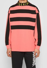 Dlouhý rukáv top v jasně růžové barvě s černými horizontálními pruhy. Obsahuje černý límec s žlutým textem "Moschino", vyrobeno z měkké tkaniny.
