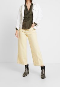 Olivengrøn omslagsbluse, hvid denimjakke, gule bukser med vide ben og sorte ankelstøvler med slangeprint og blokhæle.