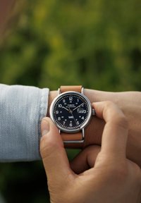 Montre ronde Timex avec un cadran noir, des index heures blancs et un bracelet en cuir marron. Aiguille réglée pour ajuster l'heure. Boîtier en acier inoxydable texturé.
