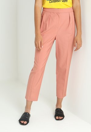 Frau trägt hoch taillierte, verkürzte pinke Hose, ein gelbes T-Shirt und schwarze offene Sandalen mit Nieten, steht vor einem weißen Hintergrund.